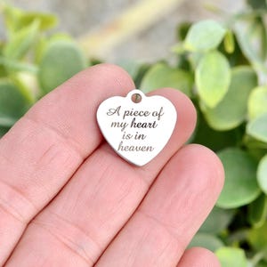 May include: A silver-toned heart-shaped metal charm featuring a small circular loop at the top. The surface is engraved with the black text A piece of my heart is in heaven, written in a cursive script font across the center of the charm.