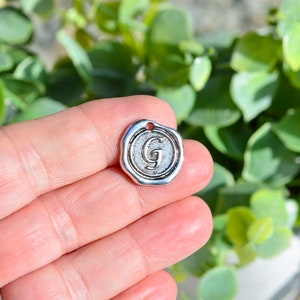 May include: Silver charm with a wax seal design featuring the letter G.