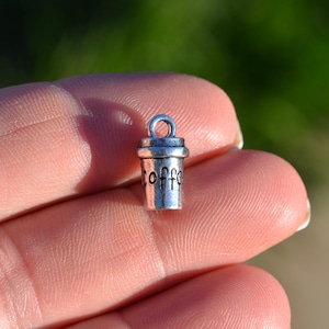May include: Silver charm in the shape of a coffee cup with the word "Coffee" printed on the side. The charm has a loop at the top for attaching to a necklace or bracelet.