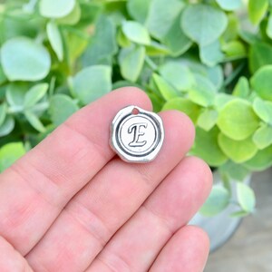 May include: Silver charm with a wax seal design featuring the letter "E" in a cursive font.