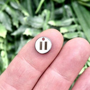 May include: A small, circular, silver-toned metal charm featuring a centered pause symbol consisting of two vertical rectangular bars and a small hole at the top for attachment, presented as a flat, polished disc with a smooth metallic finish.