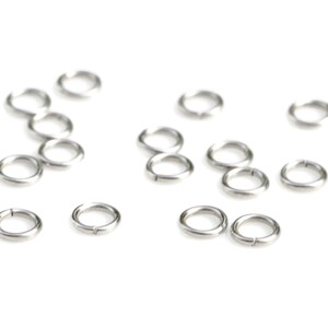 May include: A group of silver jump rings, also known as split rings, are scattered on a white background. These rings are used for connecting jewelry components.