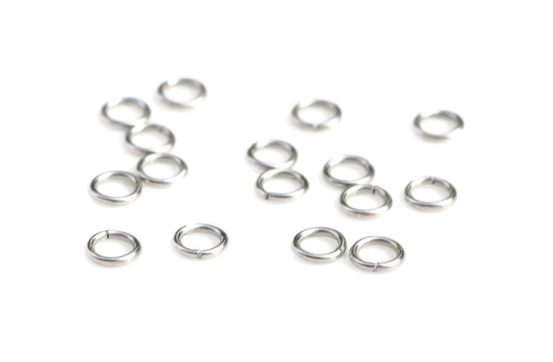 May include: A group of silver jump rings, also known as split rings, are scattered on a white background. These rings are used for connecting jewelry components.