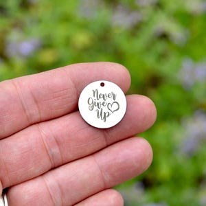 May include: A circular silver-toned metal charm featuring a small hole at the top for attachment. The surface is engraved with the black text Never Give Up, accompanied by a small heart symbol positioned to the right of the word Give.