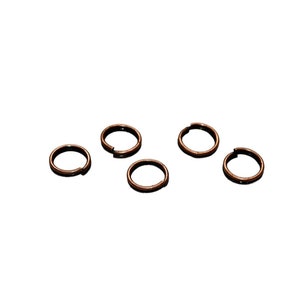 May include: Six small, round, copper-colored jump rings. The rings are all the same size and are arranged in a loose cluster.