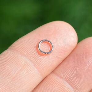 May include: A small, rose gold jump ring, held between two fingers.