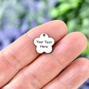 May include: A small, silver-colored metal charm shaped like a five-petaled flower. The charm features a small circular hole at the top for attachment and has the black text "Your Text Here" engraved on the front surface of the metal piece.