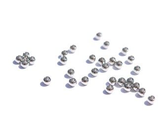 May include: A scattering of silver metal beads on a white background. The beads are small and round, and they are arranged in a random pattern.