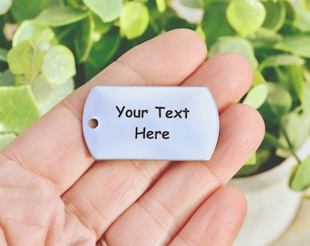 May include: A rectangular silver-toned metal dog tag with rounded corners and a single circular hole on the left side. The surface of the tag features the black printed text "Your Text Here" centred on the front of the smooth metallic plate.