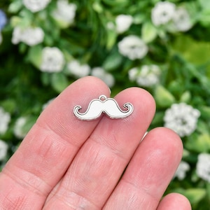 May include: A silver charm in the shape of a white mustache. The charm is being held in a person's hand.