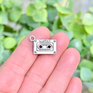 May include: A silver-toned metal charm shaped like a cassette tape. The charm has a small loop at the top for attaching to a necklace or bracelet.