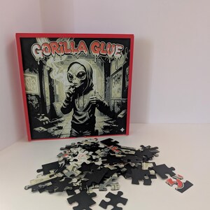 May include: A jigsaw puzzle with a red border and the words "Gorilla Glue" above an illustration of an alien smoking. The puzzle pieces are scattered in front of the box, with a few pieces assembled.