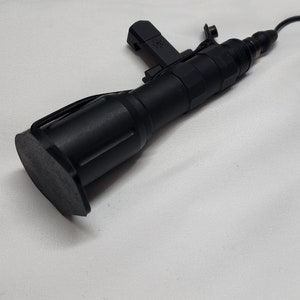 Puede incluir: Una linterna táctica negra con un interruptor de presión remoto. La linterna tiene un agarre texturizado y una tapa de extremo de goma negra. El interruptor remoto está conectado a la linterna mediante un cable negro.