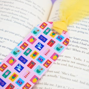May include: A pink bookmark with a repeating pattern of colorful summer-themed icons, including sunglasses, cameras, and tropical drinks. The bookmark has a yellow tassel and is placed in an open book.