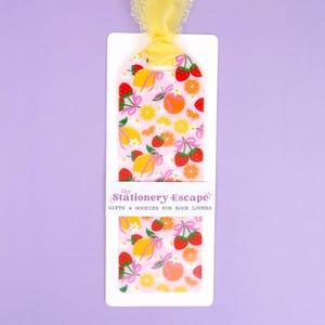 May include: A white bookmark with a pink background featuring a fruit pattern of lemons, oranges, and strawberries. The bookmark has a yellow ribbon and the text "the Stationery Escape: GIFTS & GOODIES FOR BOOK LOVERS."