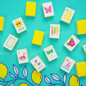 May include: A collection of rectangular mahjong tiles with white faces and yellow backs. The tiles feature colorful illustrations including a pink butterfly, a sun with sunglasses, popsicles, a pink flamingo, a dragonfly, a hibiscus, and lemons.