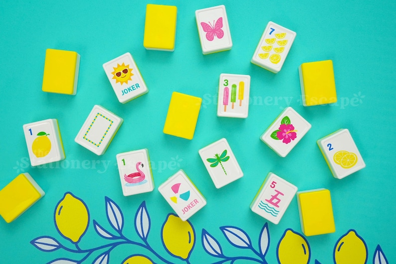 May include: A collection of rectangular mahjong tiles with white faces and yellow backs. The tiles feature colorful illustrations including a pink butterfly, a sun with sunglasses, popsicles, a pink flamingo, a dragonfly, a hibiscus, and lemons.