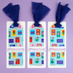 May include: Three bookmarks with a blue ribbon and a tropical stamp design. Each bookmark has a light blue background with colorful illustrations of cameras, cocktails, sunglasses, and palm trees. The text "the Stationery Escape GIFTS & GOODIES FOR BOOK LOVERS" is printed on each bookmark.