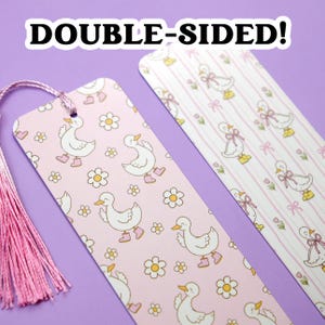 May include: Two double-sided bookmarks with a pink tassel. One bookmark features a pattern of white ducks wearing pink boots and flowers on a pink background. The other has ducks with bows on a striped background. The text "DOUBLE-SIDED!" is at the top.