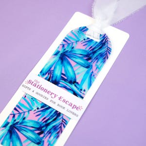 May include: A colorful bookmark featuring a tropical leaf design in shades of blue and pink. The bookmark has a white ribbon attached at the top and a white label with the text "the Stationery Escape: Gifts & Goodies for Book Lovers".