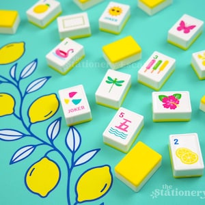 May include: A set of rectangular mahjong tiles with white faces and yellow backs scattered on a teal surface featuring a blue and yellow lemon branch illustration. The tiles display various colorful icons including a pink butterfly, a green dragonfly, a pink hibiscus, popsicles, lemons, and the text Joker.