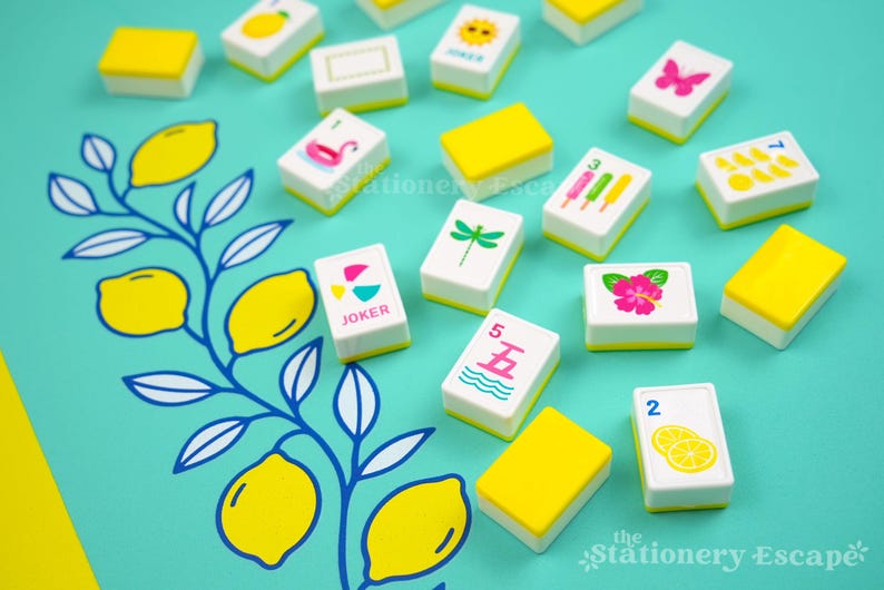 May include: A set of rectangular mahjong tiles with white faces and yellow backs scattered on a teal surface featuring a blue and yellow lemon branch illustration. The tiles display various colorful icons including a pink butterfly, a green dragonfly, a pink hibiscus, popsicles, lemons, and the text Joker.