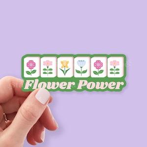 Może przedstawiać: Naklejka z zieloną obw&oacute;dką z pięcioma ilustracjami kwiat&oacute;w w kolorze r&oacute;żowym, ż&oacute;łtym i niebieskim, ze słowami "Flower Power" w kolorze r&oacute;żowym. Naklejka jest trzymana w dłoni na fioletowym tle.