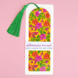 May include: A floral bookmark with a yellow background and a pattern of pink and red flowers and green leaves. The bookmark has a green tassel attached to the top. The text "the Stationery Escape" is printed on the bookmark.
