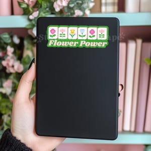 Może przedstawiać: Czarny notatnik z naklejką z napisem "Flower Power". Naklejka przedstawia rząd kolorowych kwiat&oacute;w w kolorze r&oacute;żowym, białym, ż&oacute;łtym i niebieskim. Notatnik jest trzymany w dłoni.