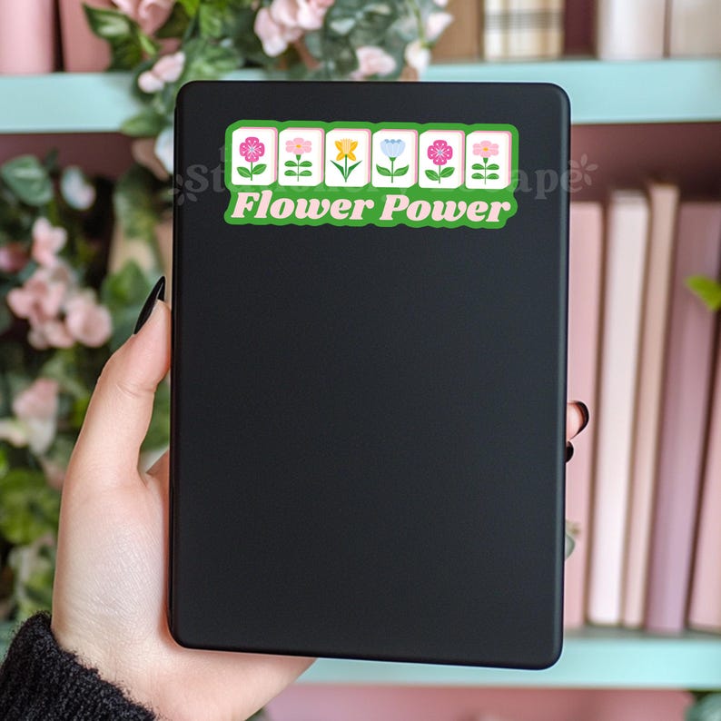 Może przedstawiać: Czarny notatnik z naklejką z napisem "Flower Power". Naklejka przedstawia rząd kolorowych kwiat&oacute;w w kolorze r&oacute;żowym, białym, ż&oacute;łtym i niebieskim. Notatnik jest trzymany w dłoni.