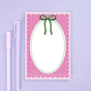 May include: A pink notepad with a scalloped edge and vertical striped pattern. A green bow adorns the top, with a white oval in the center. Two lavender pens are positioned to the left of the notepad, one with white polka dots.
