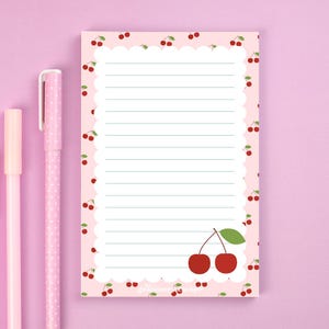 May include: A pink notepad with a scalloped edge and a cherry pattern. The notepad has lined pages for writing and two pink pens. The notepad features a pair of cherries with a green leaf in the bottom right corner.