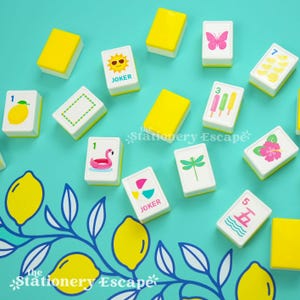 May include: A set of rectangular mahjong tiles with white faces and yellow backs, featuring colorful summer-themed illustrations including a flamingo float, popsicles, a butterfly, a sun, and lemons, scattered on a teal surface with lemon art.