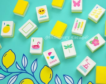 Könnte beinhalten: Ein Set rechteckiger Mahjong-Spielsteine mit weißen Vorderseiten und gelben Rückseiten, verziert mit bunten sommerlichen Motiven wie einem Flamingo-Schwimmring, Eis am Stiel, einem Schmetterling, einer Sonne und Zitronen auf türkisfarbenem Grund.
