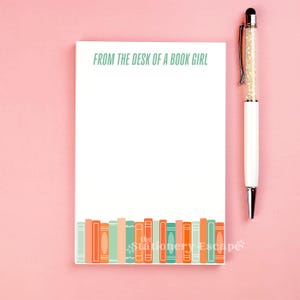 May include: A white notepad with the text "FROM THE DESK OF A BOOK GIRL" in green. The bottom of the notepad features a row of colorful book illustrations. A white pen with a crystal-filled barrel rests on a pink background.