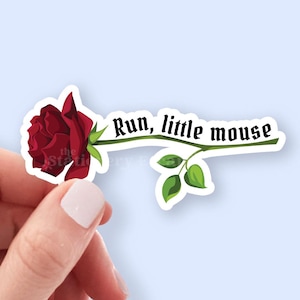 May include: A sticker featuring a dark red rose with green leaves and the text "Run, little mouse" in a whimsical font. The sticker has a white border and is held by a hand.