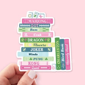 May include: A sticker featuring a stack of colorful books with titles related to Mahjong. The books are in various colors, including pink, green, and blue, with titles like "Mahjong," "Dragon," and "Joker."