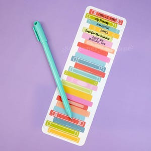 May include: A teal pen rests on a bookmark with a colorful stack of book illustrations. The bookmark features text including "FIRST LIE WINS", "My Friends", and "Just for the Summer". The background is a solid lavender color.