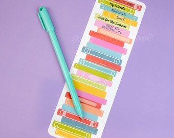 May include: A teal pen rests on a bookmark with a colorful stack of book illustrations. The bookmark features text including "FIRST LIE WINS", "My Friends", and "Just for the Summer". The background is a solid lavender color.