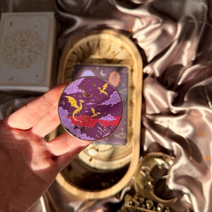 Puede incluir: Un pin esmaltado circular que muestra un cielo nocturno morado con una luna creciente dorada, estrellas y tres dragones amarillos volando. Bajo el cielo, montañas moradas estilizadas se alzan sobre un paisaje rojo, enmarcado por nubes moradas.