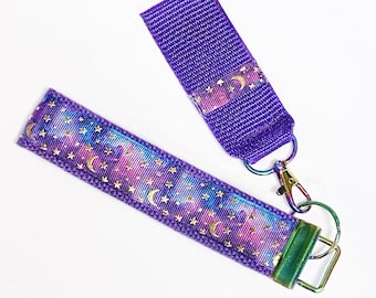 May include: A purple and iridescent key chain with a gold moon and star pattern. The key chain has a metal clasp and a purple strap with a gold moon and star pattern.