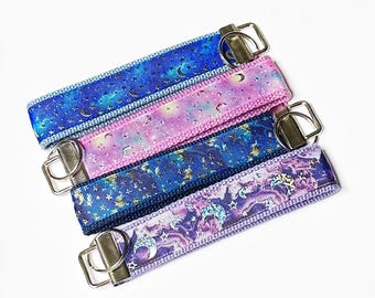 May include: Four keychains with different designs featuring a blue, purple, and pink background with gold stars and moons. The keychains are made of fabric and have a silver metal ring.