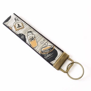 May include: A black and white fabric keychain with a witch's cauldron, potion bottles, and bubbles. The keychain has a brass key ring.