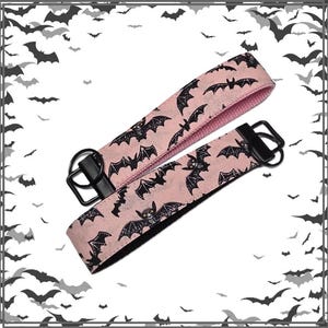 May include: Two key fobs with black hardware and a pink bat pattern. The key fobs are made of fabric and have a black metal ring. The bats are black and the background is pink.