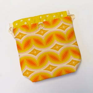 May include: A drawstring bag with a yellow and orange geometric pattern. The bag has a white lining and a silver ring at the top.