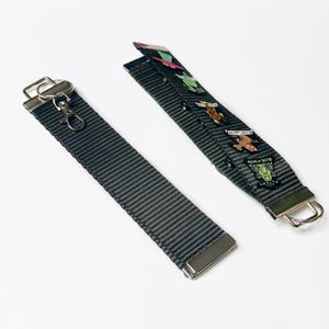May include: A gray key chain with a silver clasp and a silver ring. The key chain has a series of colorful enamel pins attached to it. The pins feature various designs, including a frog, a cat, and a mushroom.