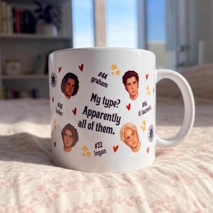 Puede incluir: Una taza de cerámica blanca con el texto "My type? Apparently all of them." rodeada de cuatro retratos masculinos ilustrados etiquetados como "#44 graham", "#46 tucker", "#66 di laurentis" y "#22 logan", decorada con pequeños corazones rojos y estrellas amarillas.