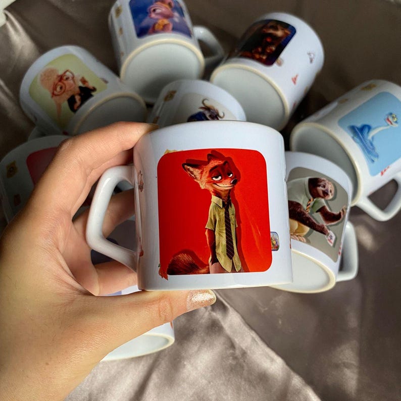 May include: A white ceramic mug featuring a graphic print of a fox character wearing a light green shirt and a dark tie against a vibrant red background. The mug has a curved handle and is part of a collection of similar mugs with various character designs.