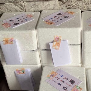May include: A stack of white rectangular polystyrene foam blocks. Each block features a white paper card attached with decorative adhesive tape. Some cards display a grid of small colorful character illustrations, while others are blank white folded cards.