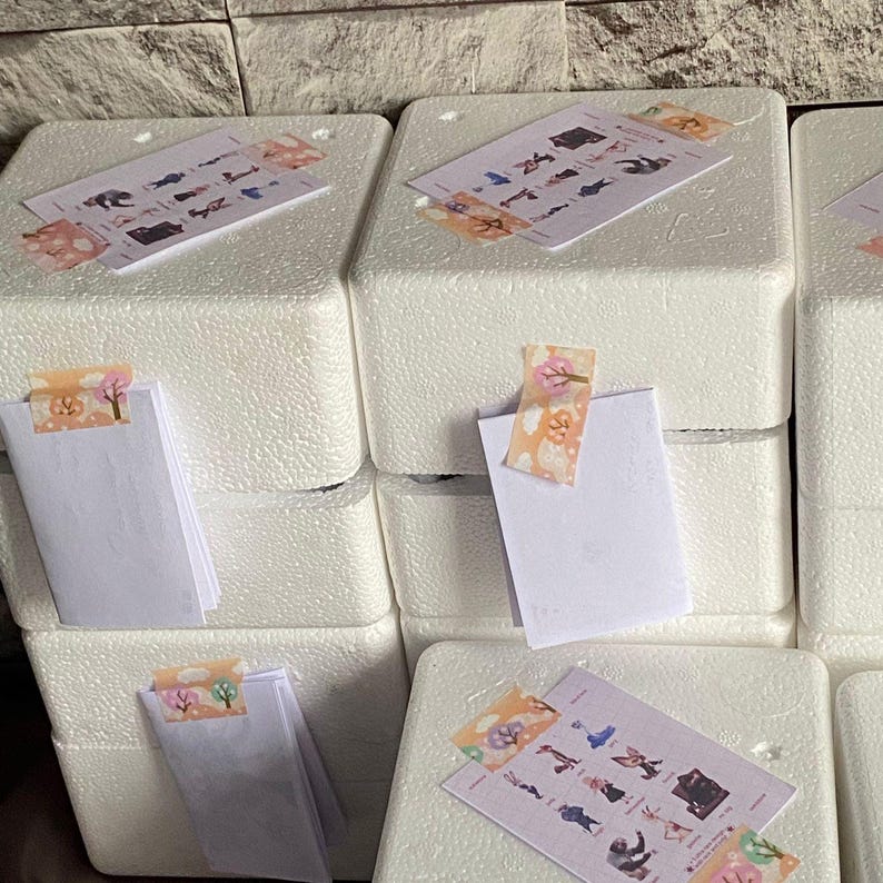 May include: A stack of white rectangular polystyrene foam blocks. Each block features a white paper card attached with decorative adhesive tape. Some cards display a grid of small colorful character illustrations, while others are blank white folded cards.